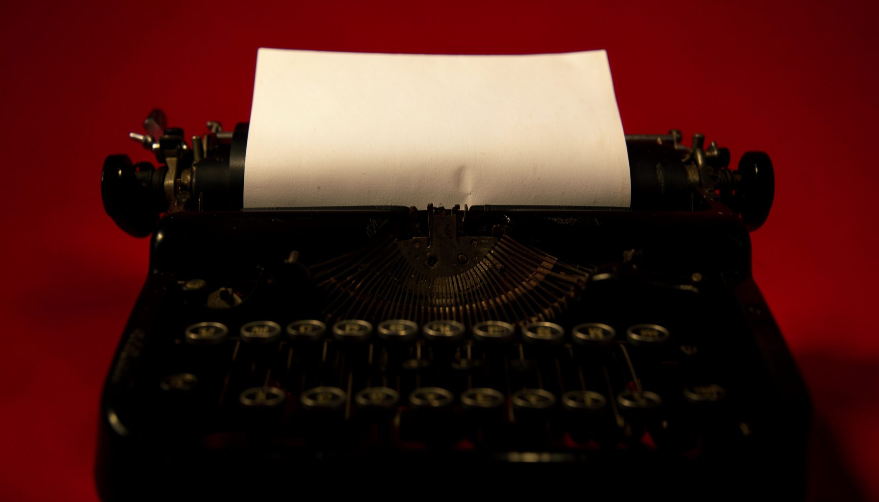 Darkened close-up of a vintage typewriter with a blank page, warm tungsten light casting deep shadows across the keys and platen, moody and contemplative atmosphere.